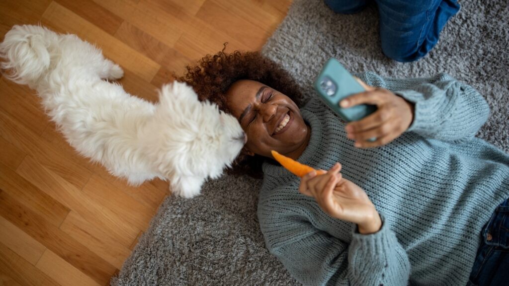 A lady with her eyes closed lying on the floor laughing and looking at her phone whilst holding a carrot next to a small white dog