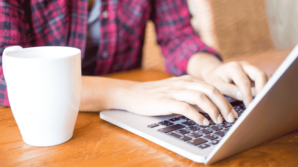 Person's hands typing on a laptop next to a white mug