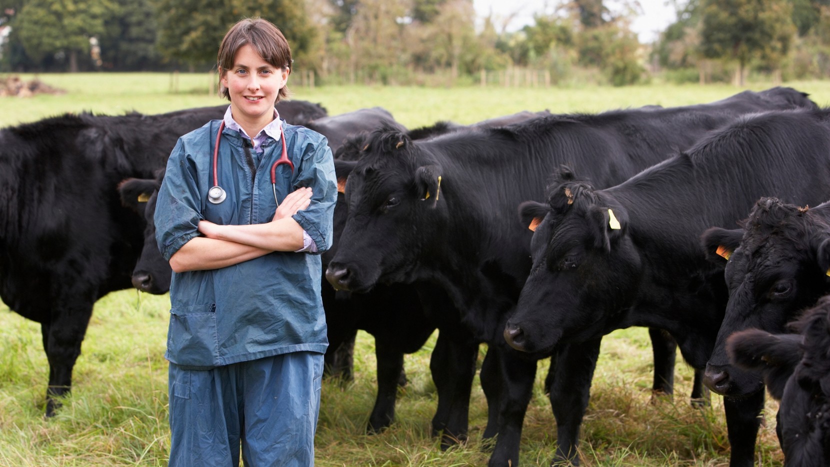 A smiling vet standing in front of a herd of cows in a grassy field with her arms crossed