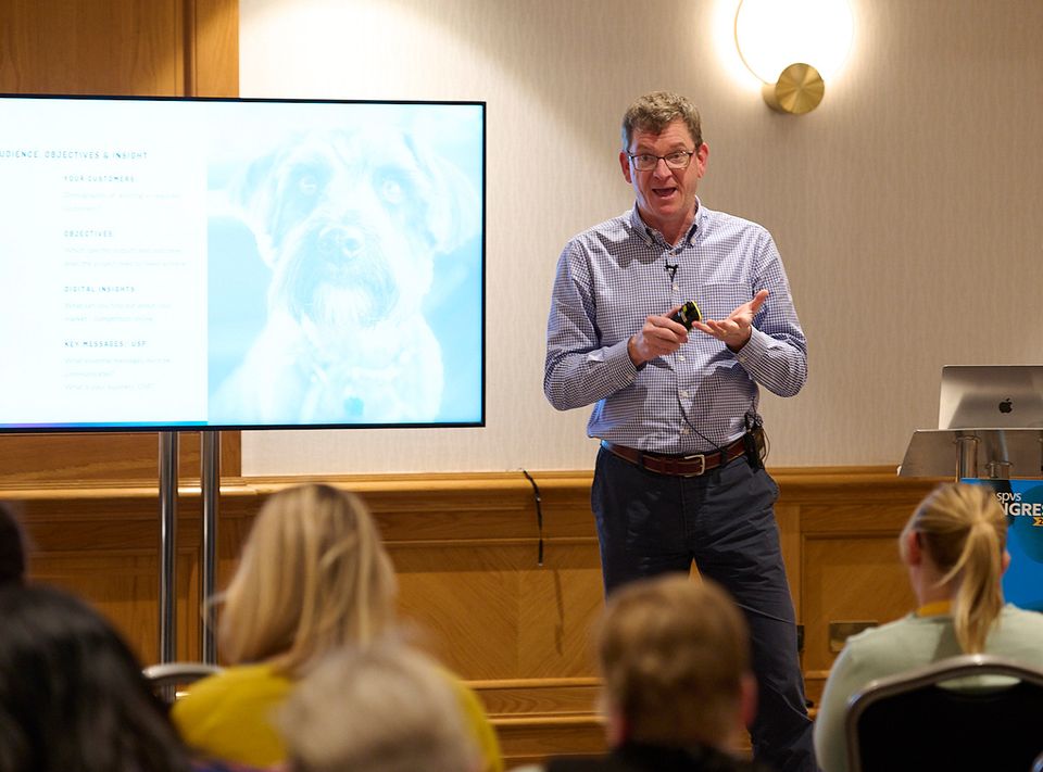 Andrew Rastall is standing and giving a talk in a conference room with a large screen in front of an audience